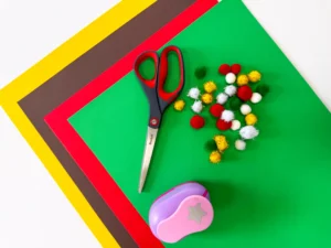 supplies for this handprint tree christmas card: Scissors resting on bright layered craft papers with colorful pom-poms scattered nearby, suggesting a DIY craft setup.