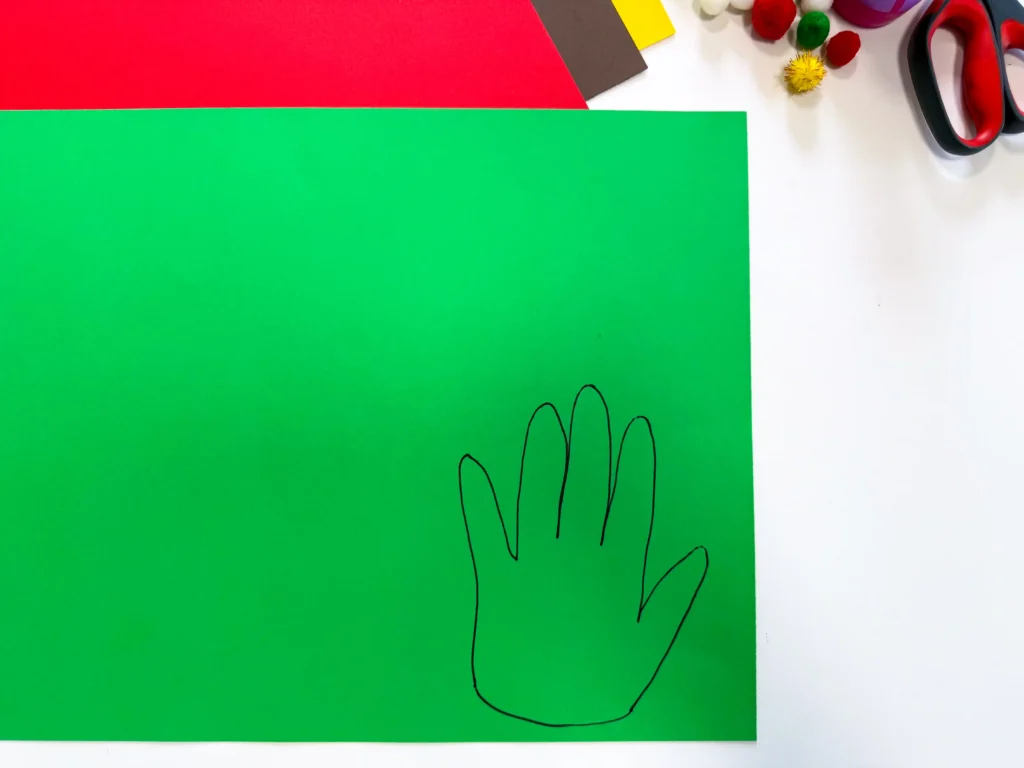 Bright green craft paper with a simple black hand outline; red and yellow paper scraps and scissors sit on a white desk to the side, suggesting an arts-and-crafts scene.