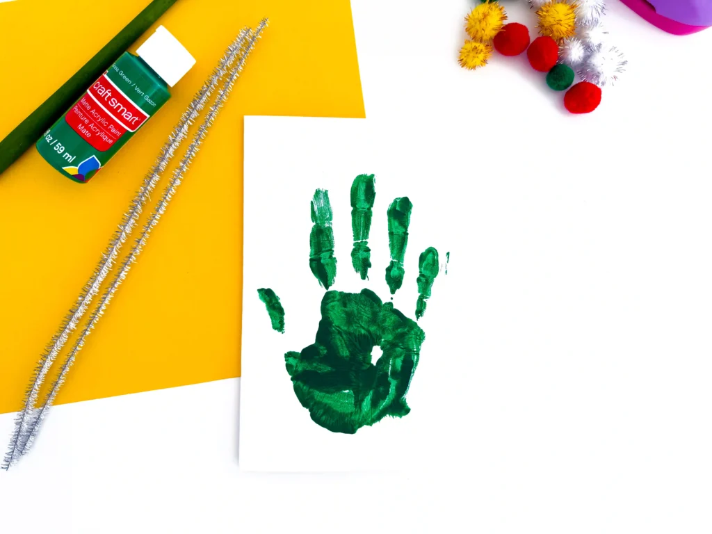 Green handprint on white paper with craft supplies: glittery pipe cleaners, colorful pom-poms, and a green acrylic paint bottle on a yellow background.