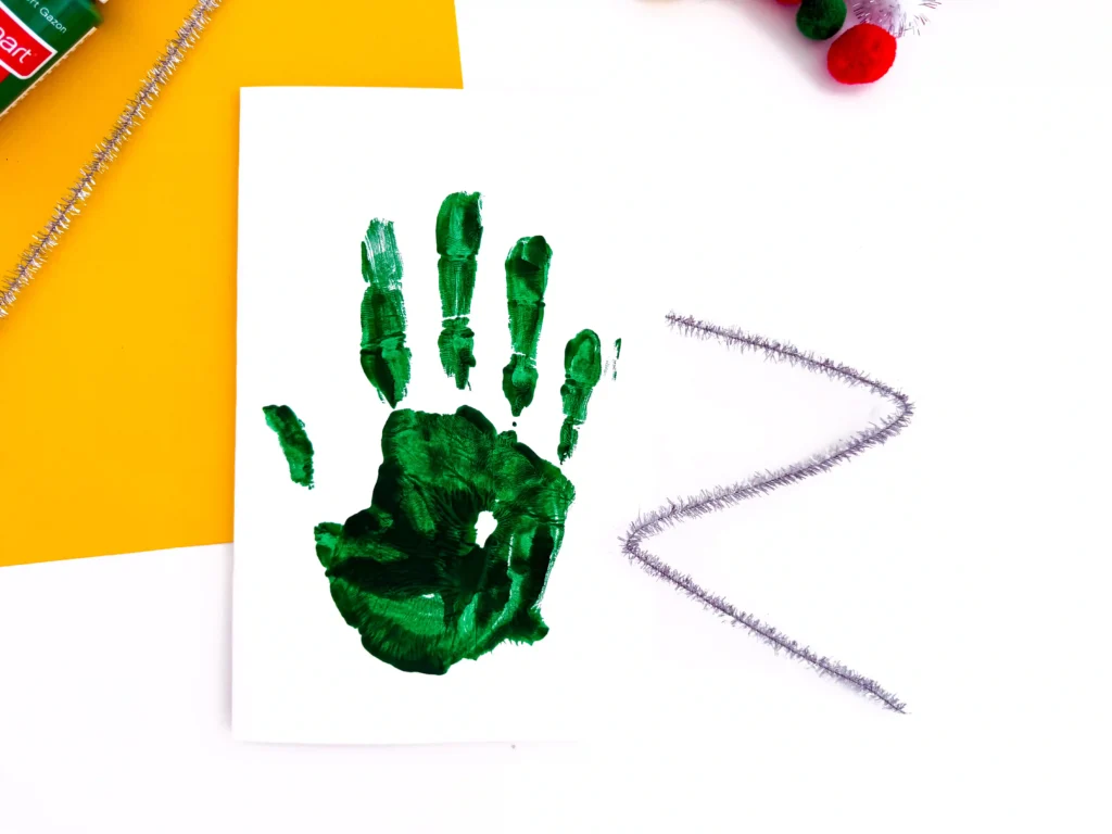 Green paint handprint on a white sheet of paper and a z shaped silver glittery pipe cleaner next to it