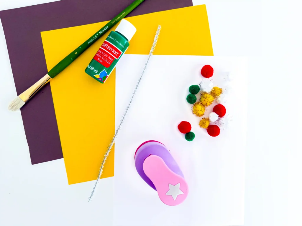 supplies for this handprint christmas tree card: green paint bottle, two silver tinsel pipe cleaners, and blank white cards with a colorful pompom garland in the corner