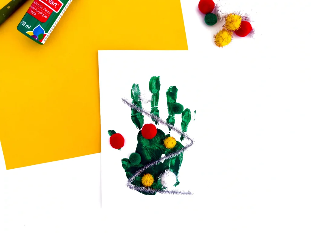 Craft scene: white sheet with a green painted handprint and multicolored pom-poms on a bright yellow background.