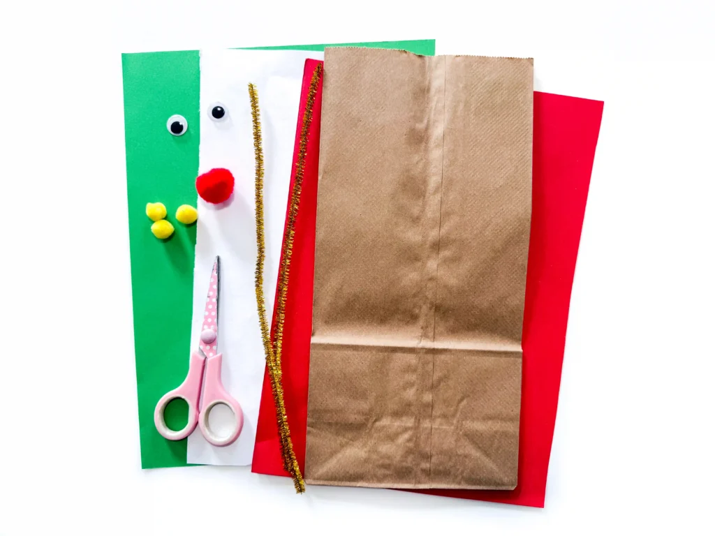 supplies for the reindeer paper bag craft: green, white, and red paper, brown paper bags, scissors, gold pipe cleaners, pom poms, and googly eyes ready for a DIY project.