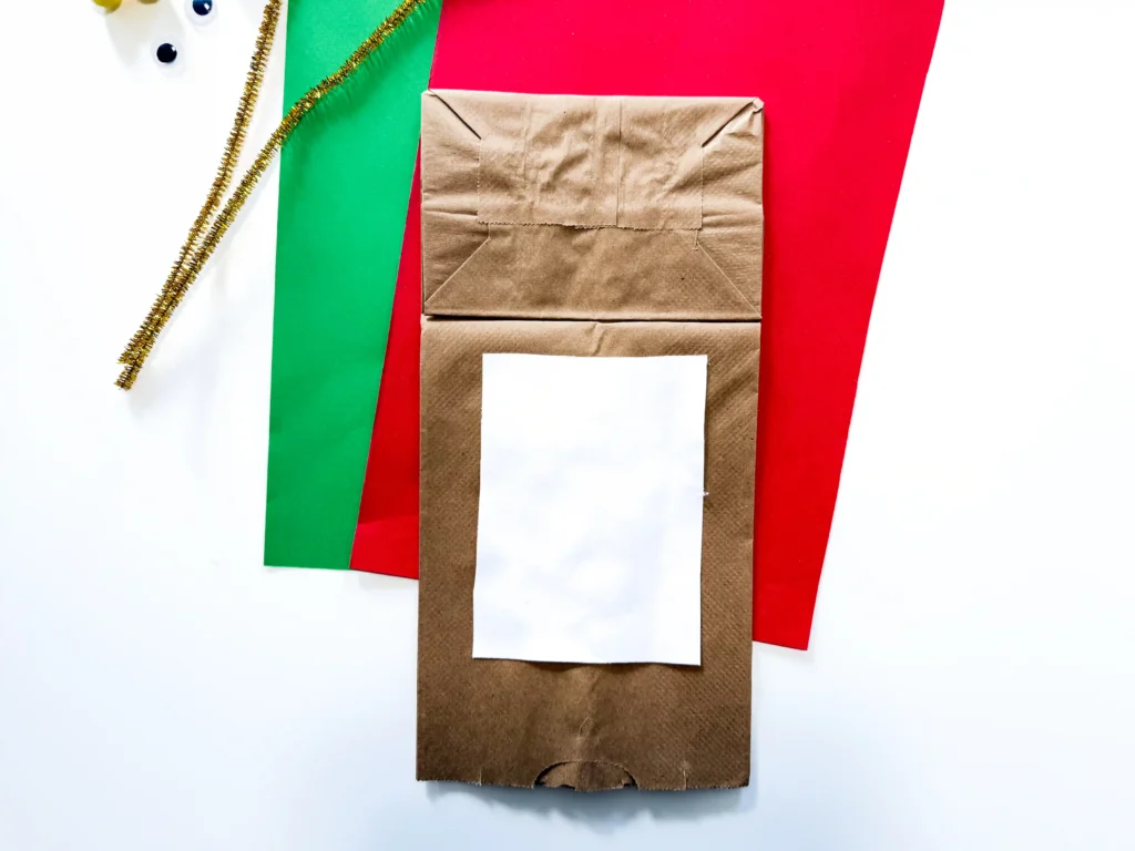 Brown paper gift bag with a white label sits on red and green paper backgrounds with a gold tinsel garland to the left.