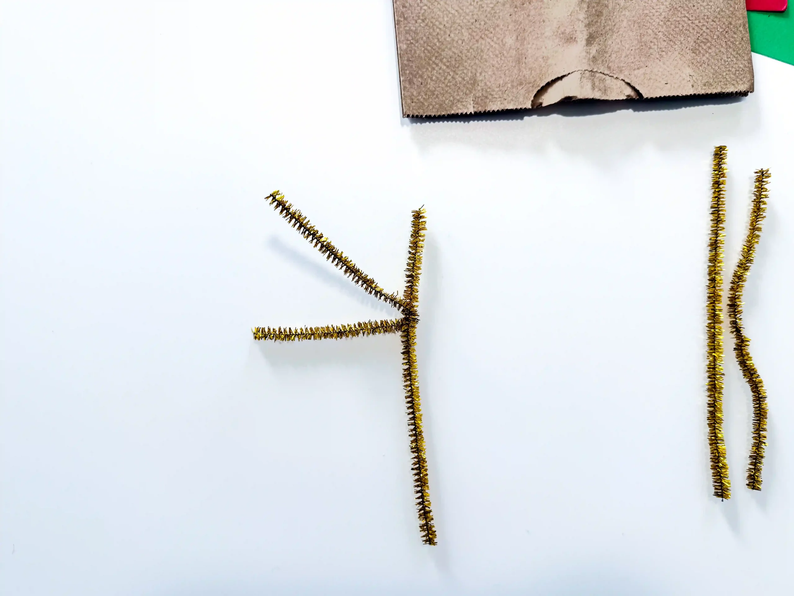 White background with gold pipe-cleaner stems arranged in a branching, vertical pattern and a brown cardboard piece at the top edge.