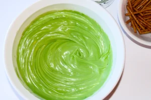 Bright green swirled frosting in a white bowl, with pretzel sticks on a plate to the right.