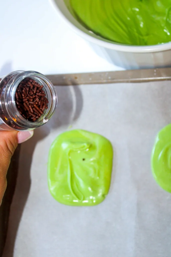 Hand holding a glass jar of small brown sprinkles next to bright green fondant shapes on a baking sheet.