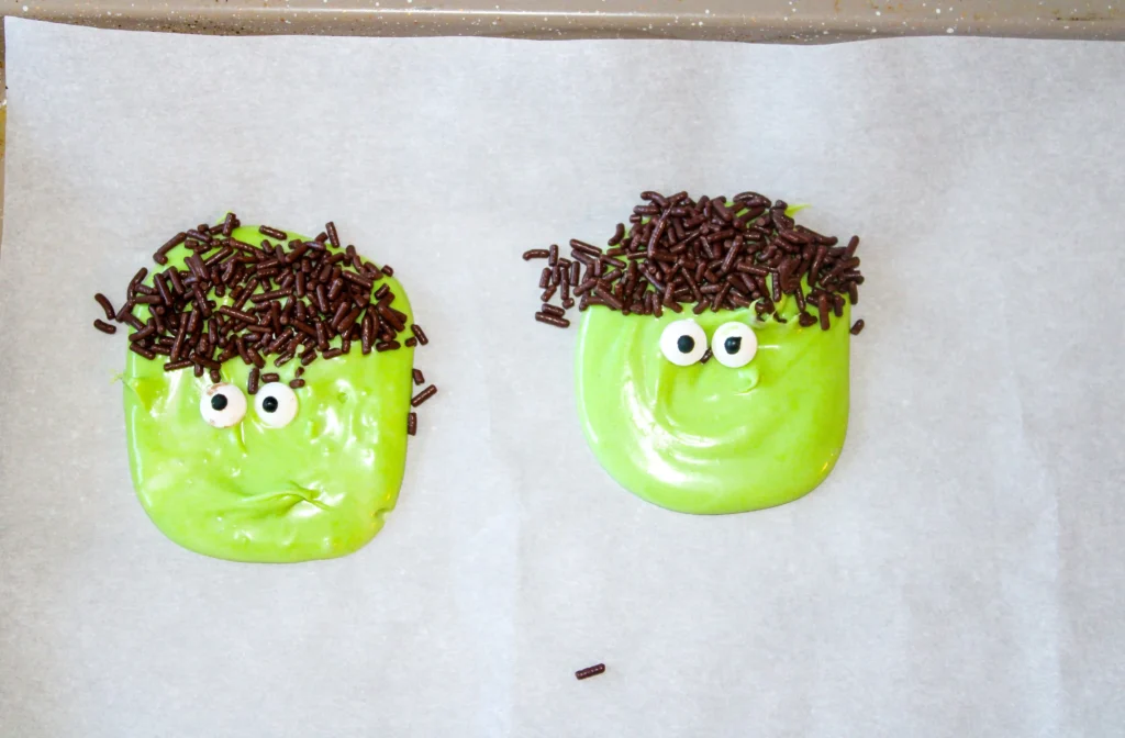 Two green pieces of white chocolate bark with brown sprinkles and googly eyes on parchment paper.