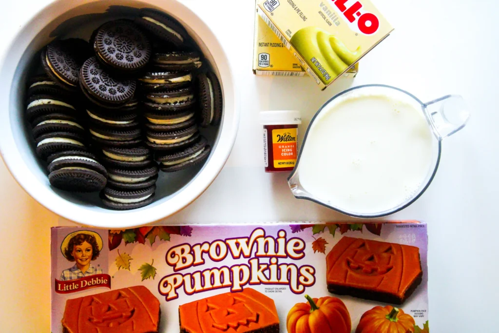 ingredients for the halloween dirt cups: a bowl full of chocolate sandwich cookies (Oreos), a carton of milk, a box of Little Debbie brownie pumpkins mix, and a bottle of Wilton orange icing color.