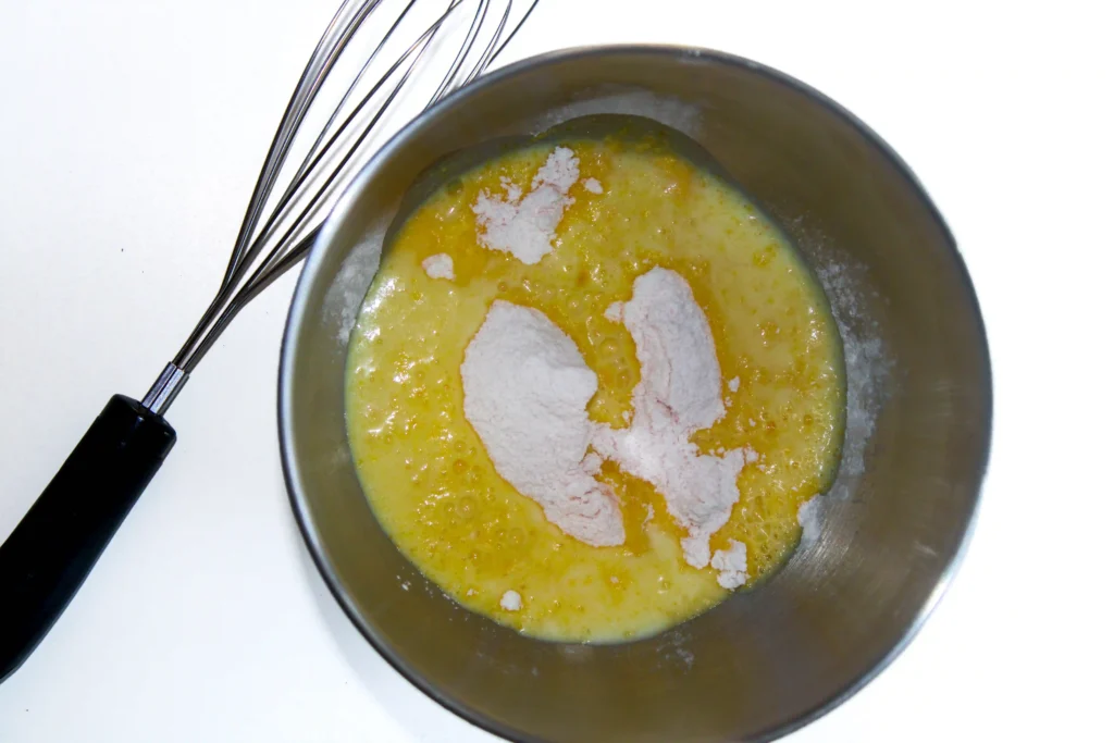 Metal mixing bowl with yellow batter and white flour sprinkled on top; a whisk with a black-handled grip rests nearby on the white surface.