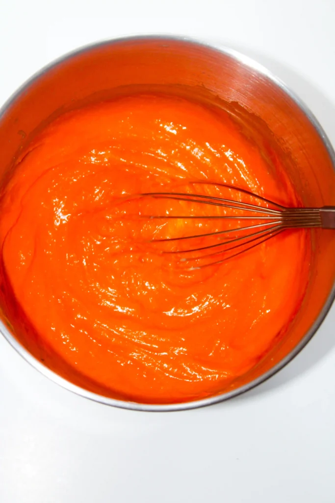 Close-up of bright orange icing in a metal mixing bowl with a whisk resting in it.