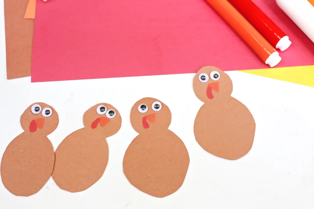Row of brown paper turkey shapes with googly eyes and orange beaks on white craft paper, with pink and red construction paper and colored pencils in the background.