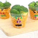 Three clear cups on a wooden surface, each filled with pasta and topped with broccoli florets, decorated with colorful paper shapes for a playful snack.