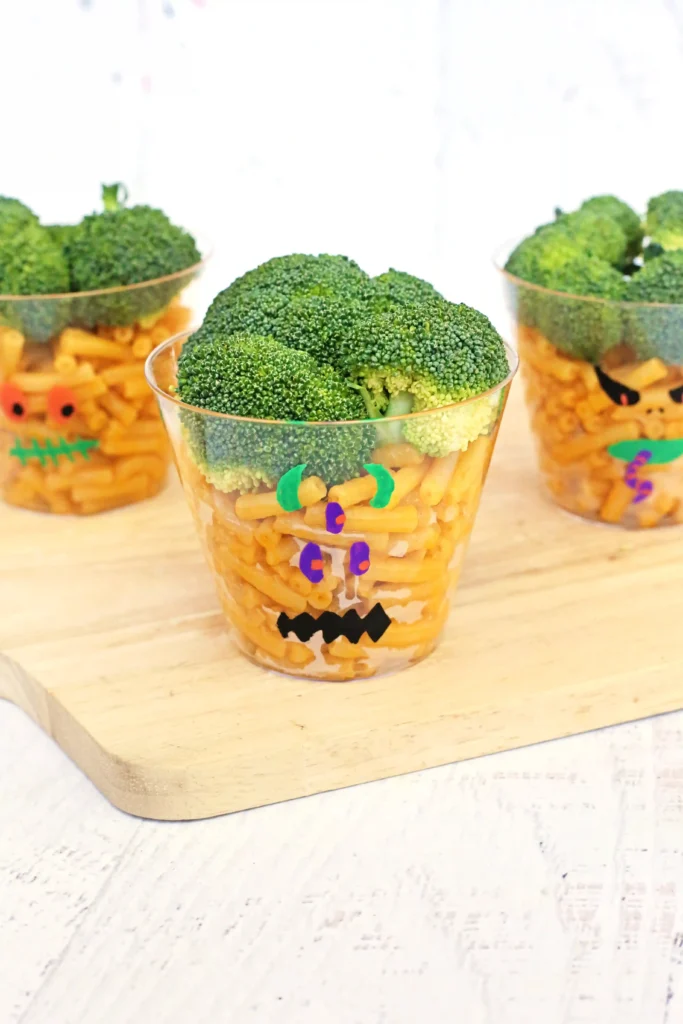 Three clear cups on a wooden surface, each filled with pasta and topped with broccoli florets, decorated with monster faces