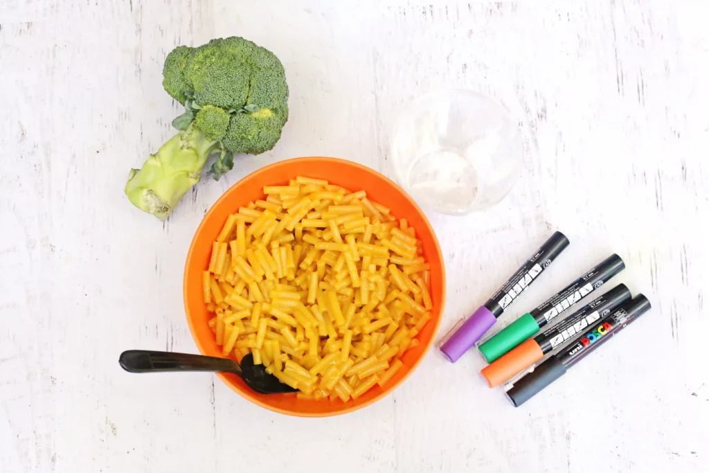 ingredients for the monster mac and cheese: Top-down view of an orange bowl filled with macaroni and cheese, with a broccoli floret beside it on a white wooden table, and marker pens nearby.