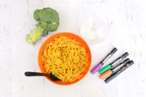 ingredients for the monster mac and cheese: Top-down view of an orange bowl filled with macaroni and cheese, with a broccoli floret beside it on a white wooden table, and marker pens nearby.