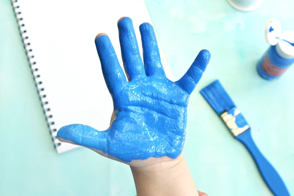 Child's hand covered in bright blue paint resting on a craft table.