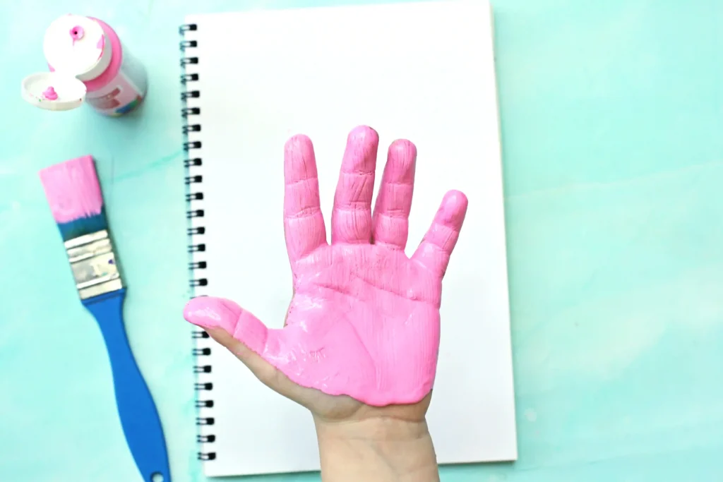 Top-down view of a pink-painted hand raised over a white spiral notebook, with a pink paint bottle and blue brush on a turquoise background.