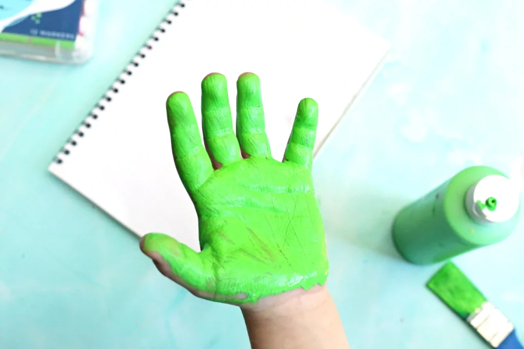Bright green-painted hand with fingers spread, on a light blue surface next to a notebook and crafting supplies.