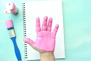 Top-down view of a pink-painted hand raised over a white spiral notebook, with a pink paint bottle and blue brush on a turquoise background.