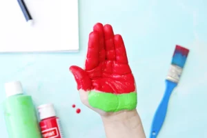 Hand raised with red-painted fingers and a green-painted wrist, art supplies visible in a light blue workspace background.