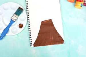 Blue-handled paintbrush rests on a white paint palette with brown paint; a spiral notebook sits open with brown strokes on its page against a turquoise surface.