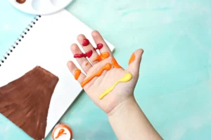 Hand covered in orange, red, and yellow paint, with a notebook and paint pot visible in the background.