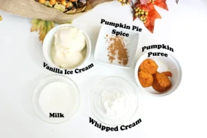 ingredients for the pumpkin pie milkshake: vanilla ice cream, whipped cream, milk, pumpkin puree, and pumpkin pie spice in white bowls, with autumn leaves in the background. The image also shows overlaid text labels such as 'Vanilla Ice Cream', 'Milk', 'Whipped Cream', 'Pumpkin Puree', and 'Pumpkin Pie Spice'.