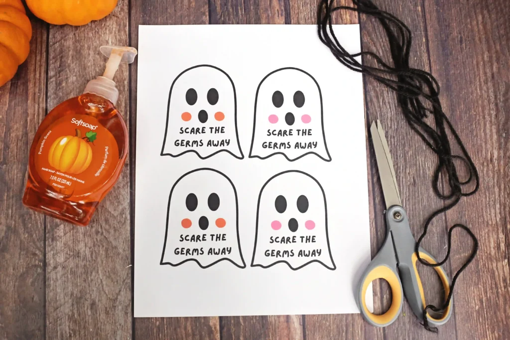 Printable sheet featuring four ghost illustrations with the caption'Scare the germs away' on a wooden table, alongside Halloween soap, scissors, and string strands.