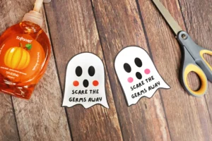 Orange Softsoap hand soap bottle on a wooden surface with three ghost-shaped tags that say 'Scare the germs away' and a pair of scissors nearby.
