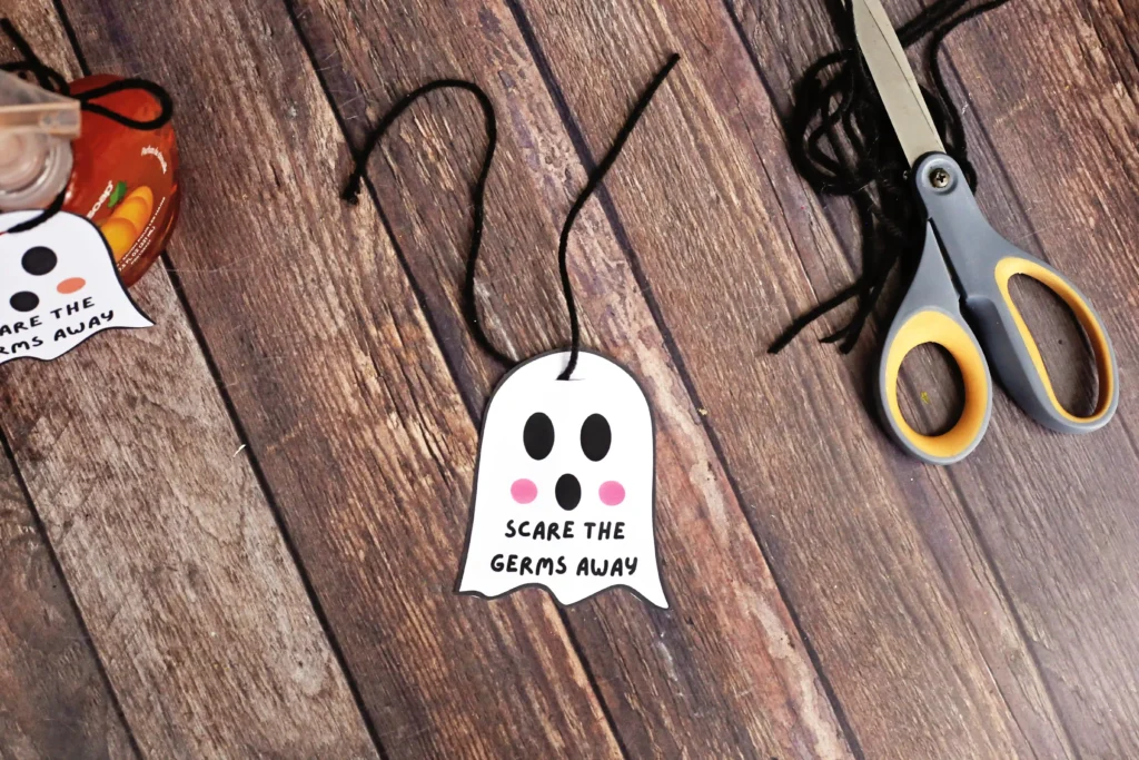 Ghost-shaped tag with black eyes and pink cheeks reading'SCARE THE GERMS AWAY' on a string, placed on a wooden table near scissors and a jar of jam.