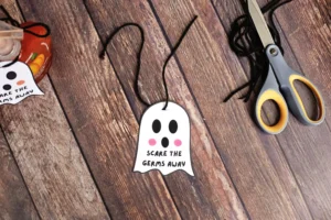 Ghost-shaped tag with black eyes and pink cheeks reading 'SCARE THE GERMS AWAY' on a string, placed on a wooden table near scissors and a jar of jam.