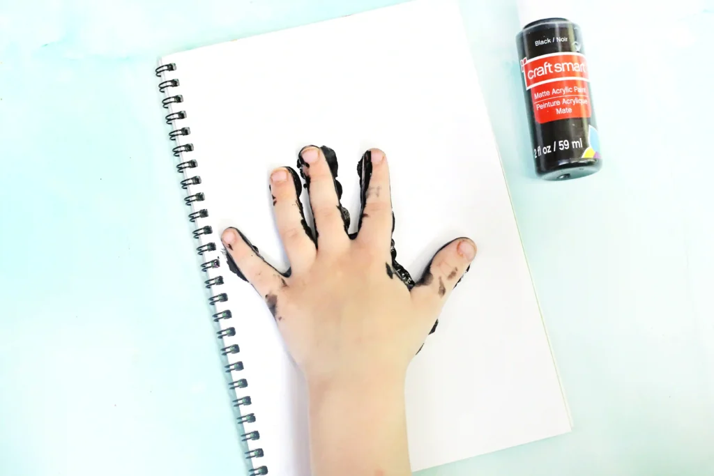 Open spiral notebook with a hand stained in black paint resting on a page, and a bottle of black Craft Smart acrylic paint on the right side.