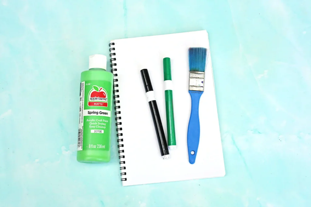 supplies for the letter t handprint craft: green paint bottle, notebook, markers, and a blue paintbrush on a white page against a light cyan background.