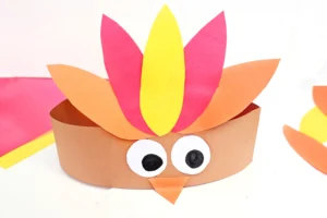 Paper craft turkey with a brown body, white and black eyes, an orange beak, and bright colored feathers in pink, yellow, and orange.