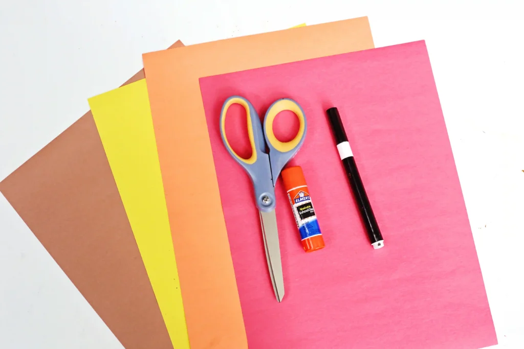supplies for the turkey headband craft: Scissors, glue stick, and a marker resting on layered colored construction paper (pink, orange, yellow, brown).