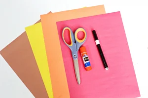 supplies for the turkey headband craft: Scissors, glue stick, and a marker resting on layered colored construction paper (pink, orange, yellow, brown).