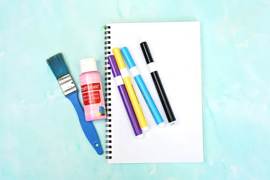 supplies for the letter u handprint craft: paintbrush, pink acrylic paint, colorful markers, and a blank page on a light blue surface.