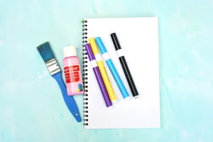 supplies for the letter u handprint craft: paintbrush, pink acrylic paint, colorful markers, and a blank page on a light blue surface.