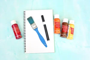 supplies for the letter v handprint craft: Blank spiral notebook on a turquoise surface with scattered acrylic paints (red, orange, brown, yellow), a blue paintbrush, and a black marker nearby.