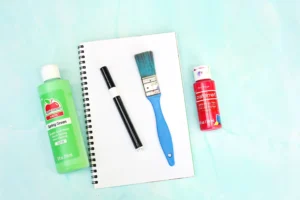 supplies for the letter w handprint craft: spiral notebook, black marker, blue paintbrush, and green and red acrylic paint bottles nearby.