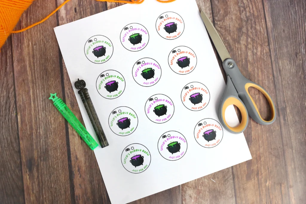 supplies for the printable Halloween tags: Sheet of circular stickers featuring a black cauldron and colorful'Witch's Bubbly Brew' text, laid on a craft paper next to scissors and pens on a wooden table.