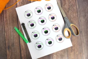 supplies for the printable Halloween tags: Sheet of circular stickers featuring a black cauldron and colorful 'Witch's Bubbly Brew' text, laid on a craft paper next to scissors and pens on a wooden table.