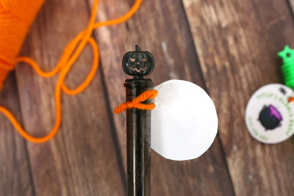 Close-up of a small black jack-o'-lantern atop a dark bubble wand, with orange string and Halloween decorations in the background.