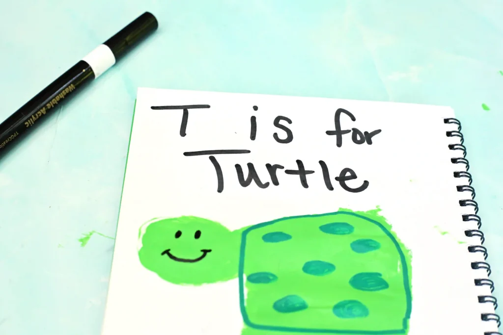 Notebook page showing a green cartoon turtle drawn with marker, with the text'T is for Turtle' handwritten above/around it, and a black marker in the corner.