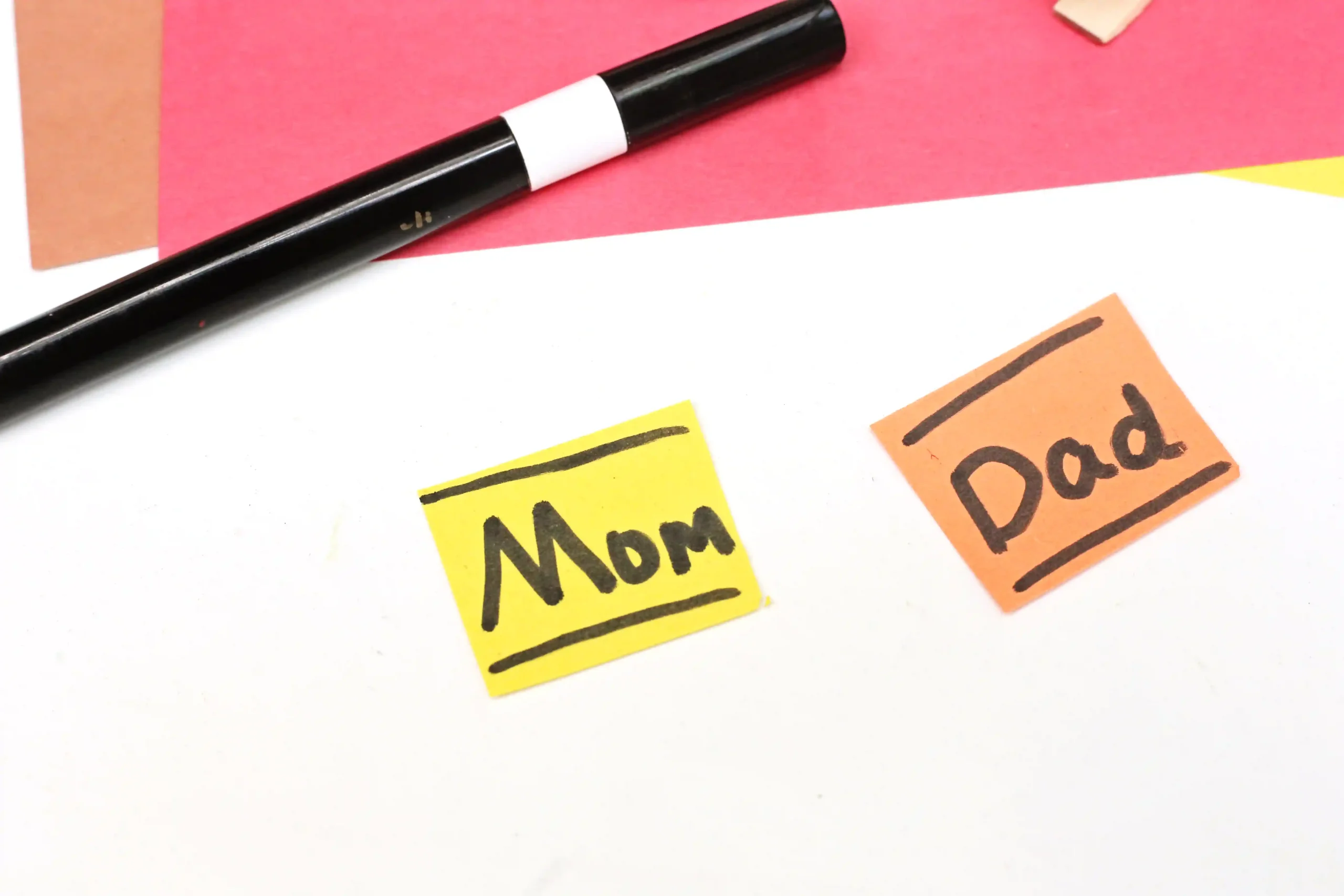 Top-down view of a black marker on red paper with yellow note saying 'Mom' and an orange note saying 'Dad' nearby.