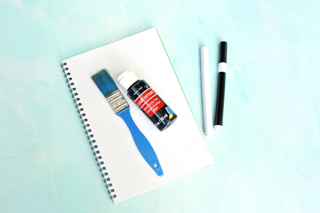 supplies for the letter x handprint craft: Spiral notebook with white pages, blue paintbrush, white and black markers on a light blue surface.