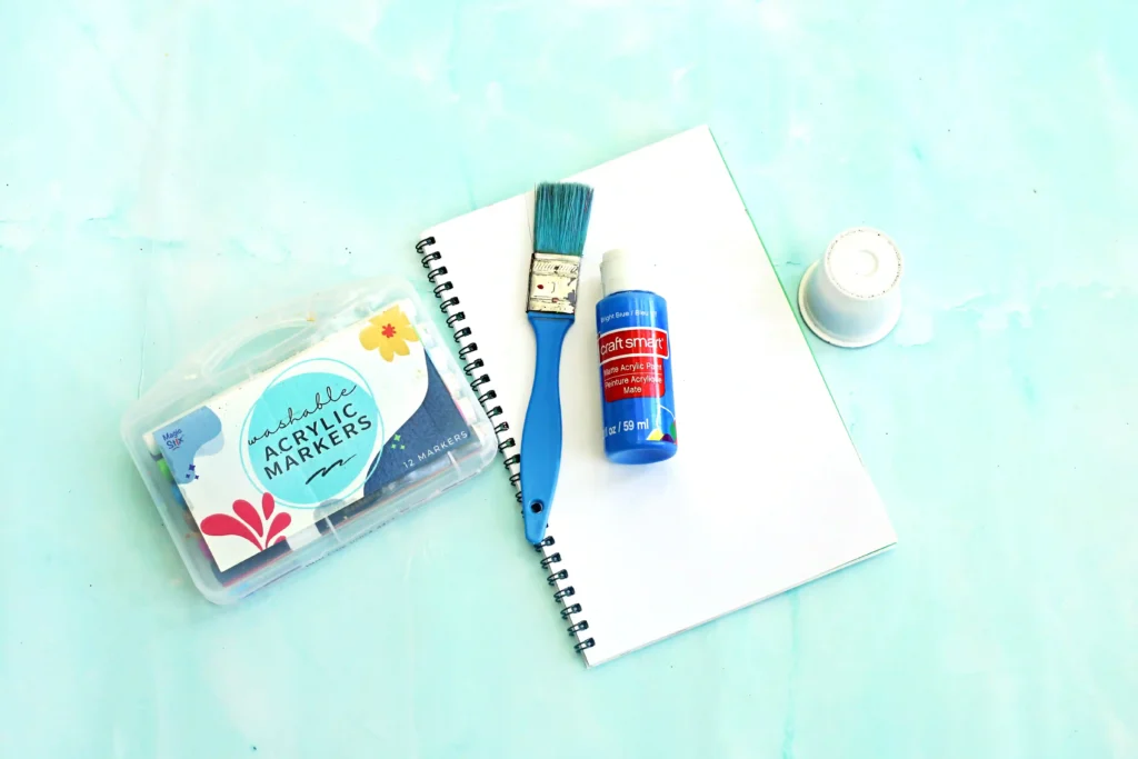 supplies for the letter y handprint craft: a blue-handled paintbrush, a blue acrylic paint bottle, and a spiral notebook on a pale blue surface with a marker set nearby.