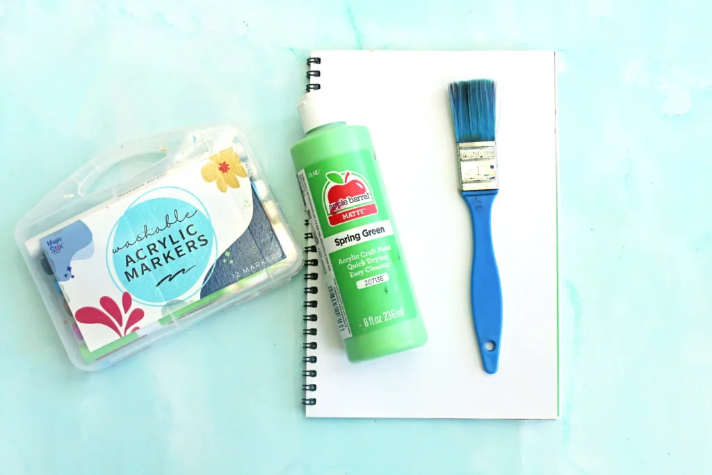 supplies for the letter z handprint craft: a notebook, a green Apple Barrel acrylic paint bottle, a blue paintbrush, and a case of Washable Acrylic Markers.