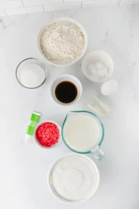 ingredients for the grinch pancakes: flour in a bowl, sugar, vanilla, eggs, milk, pink heart candies, and icing tube on a marble surface.